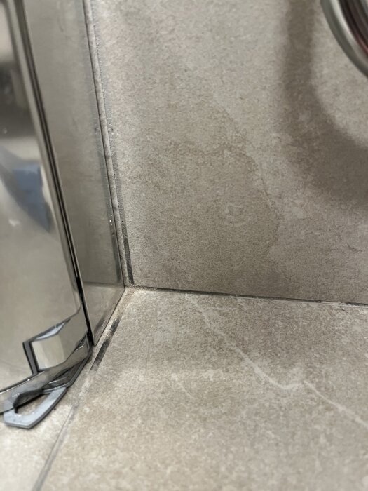 Närbild av ojämna badrumsplattor med ojämn fog och synliga skador på vissa plattor nära duschkabinens kant.