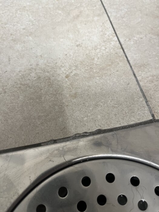 Närbild av ojämnt lagda kakelplattor i badrum nära ett duschavlopp. Plattorna är osymmetriska och fogen ser flammig ut.