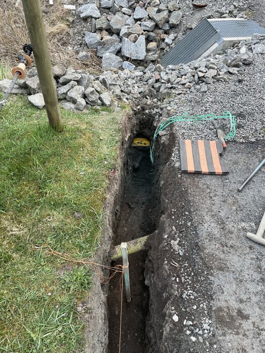 Grävd grop i gräsmattan med ett dräneringsrör som leder mot en stenkista. Stolpe synlig, omgiven av stenar och grus vid sidan.