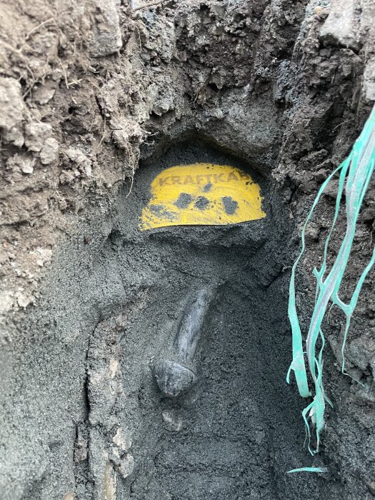 Uppgrävd kabel i en grävd utgrävning, markerad med en gul skylt där det står "kraftkabel", omgiven av jord och rötter.