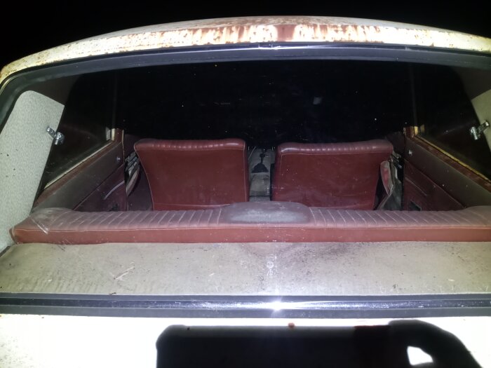 Rostig tvådörrarsbil sedd bakifrån, med bruna säten och instrumentpanel, sparsamt baksäte och mörk omgivning.