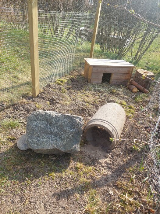 En trädgårdsinhägnad för kaniner med ett litet kaninhus, en stor sten och ett betongrör på marken.