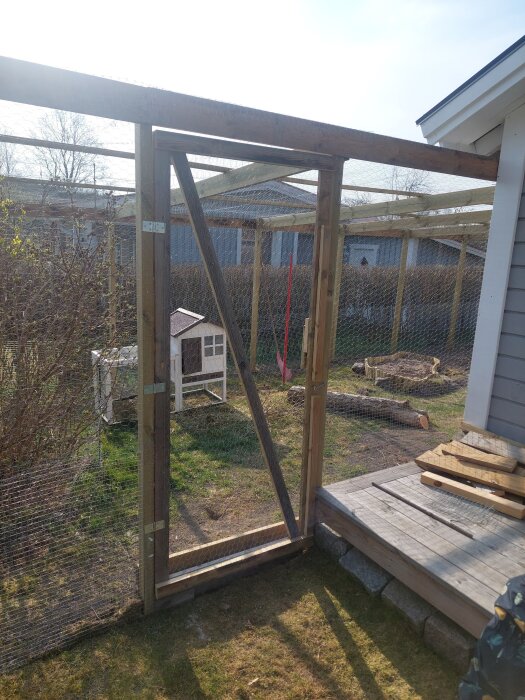 Utomhuskaninbur i trä och nät intill trädäck, inuti syns ett litet vitt kaninhus.