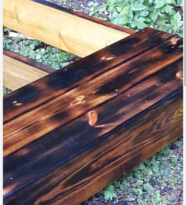 Brända träbrädor placerade på marken, sannolikt för att användas till odlingslådor, med växter i bakgrunden.