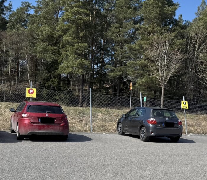 Två parkerade bilar på en parkeringsplats framför ett skogsområde, med skyltar för parkering och elbilsladdning i bakgrunden.