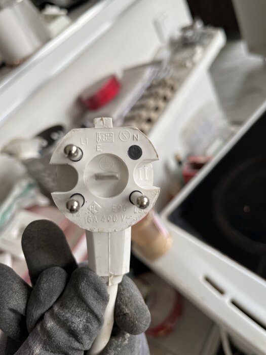 En hand håller i en Perilex-stickpropp märkt med 16A 400V, mot en oskarp bakgrund som verkar vara en arbetsyta eller bänkskiva.