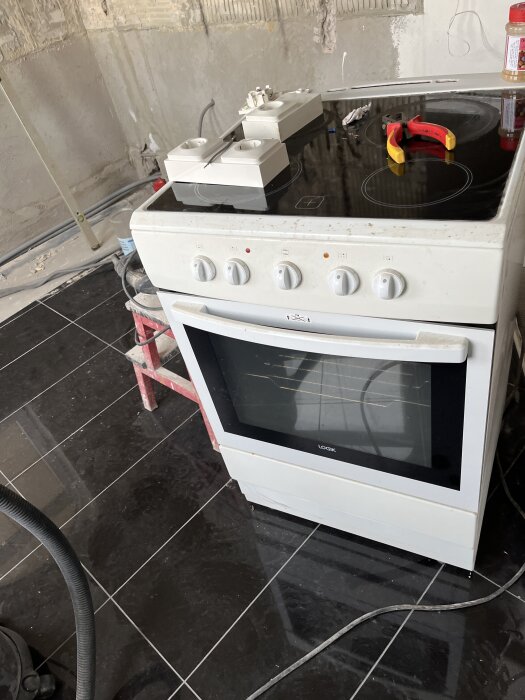 Vit spis med keramikhäll, liggande verktyg och eluttag ovanpå, placerad på svart kakelgolv i ett halvfärdigt kök.