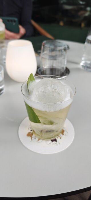 En cocktail med en stor rund iskula och ett grönt blad i glaset, placerad på ett bord bredvid ett stearinljus.