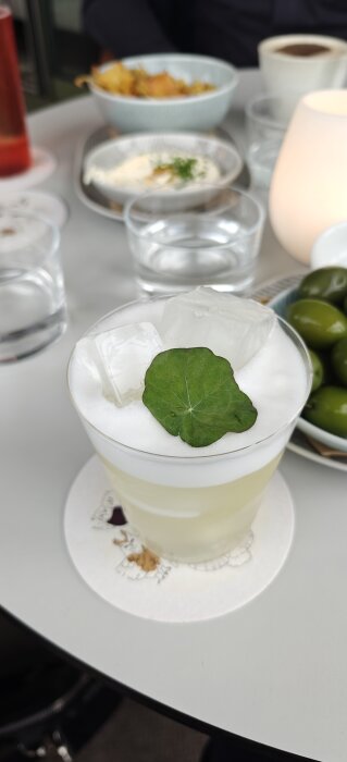 Banandrink med skum och isbitar, toppad med ett grönt blad, serverad på ett bord bredvid oliver och andra tilltugg.