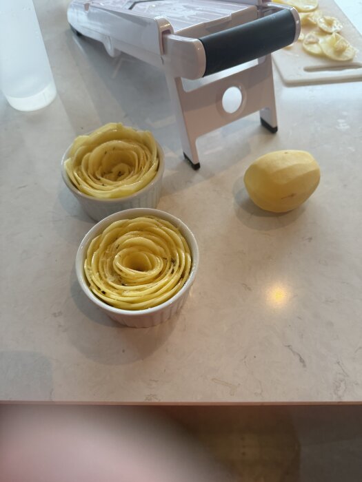 Potatisrosor i små ramekinformar på ett köksbord, bredvid en skalad potatis och en mandolin.