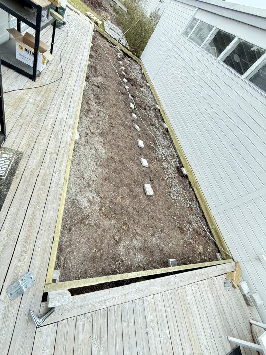 Byggarbete för altan med trästomme och linjer av stenar på marken bredvid ett hus; befintlig altan i förgrunden.