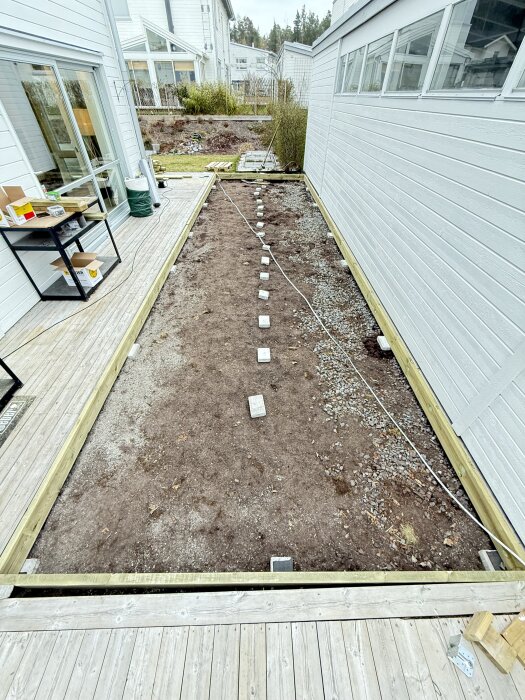 Ramkonstruktion för altanutbyggnad vid sidan av ett hus, med markstenar och rep utlagda på marken, synligt befintligt trägolv i förgrunden.