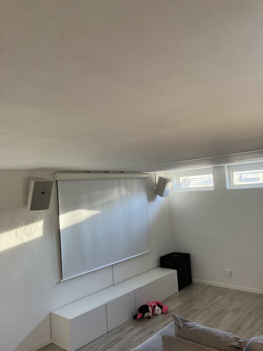 Hemmabio i källare med projektorduk, ljudhögtalare, en leksak på golvet och soffa i vitt rum.