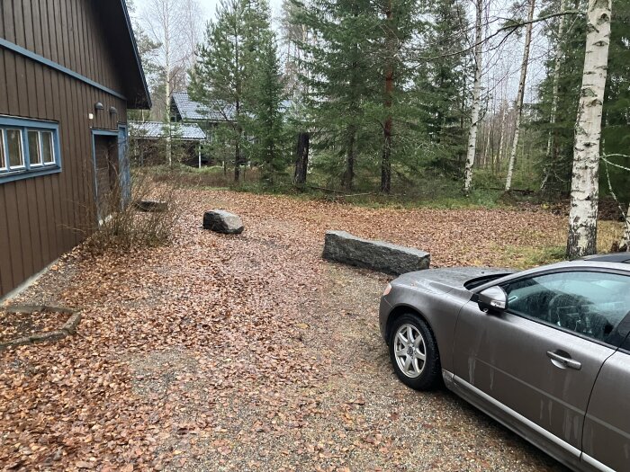 Garageuppfart med stenar vid kanten, silverfärgad bil till höger och omgiven av träd och löv.
