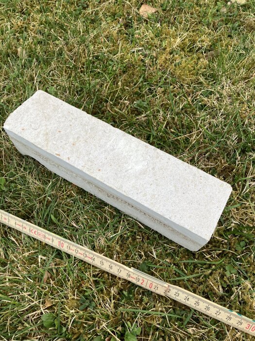 En vit tegelsten ligger på gräsmatta med måttband bredvid.