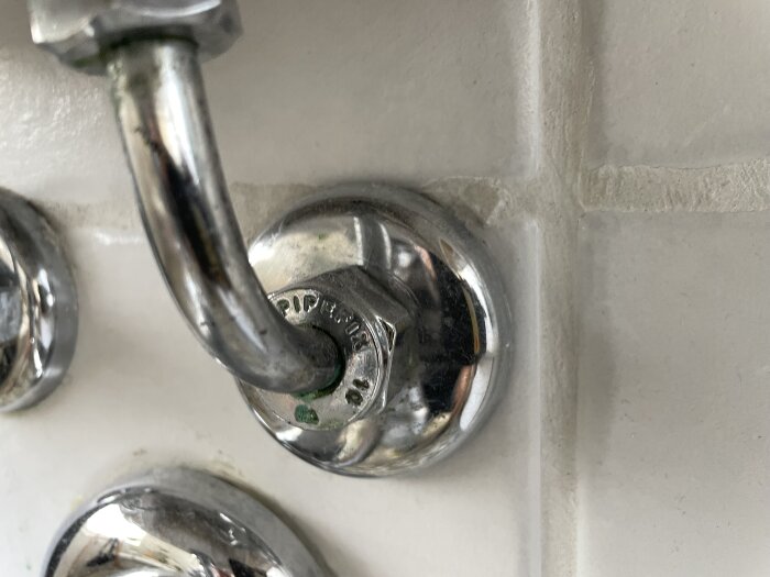 Kromad pipventil installerad på vitt kakel i ett trångt utrymme bakom ett handfat.