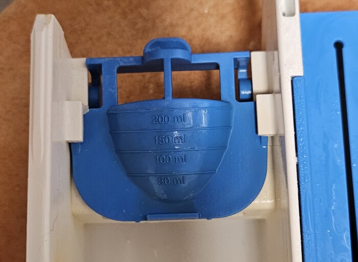 Blå tvättmedelslucka i öppet läge med måttmarkeringar 50-200 ml för flytande tvättmedel.