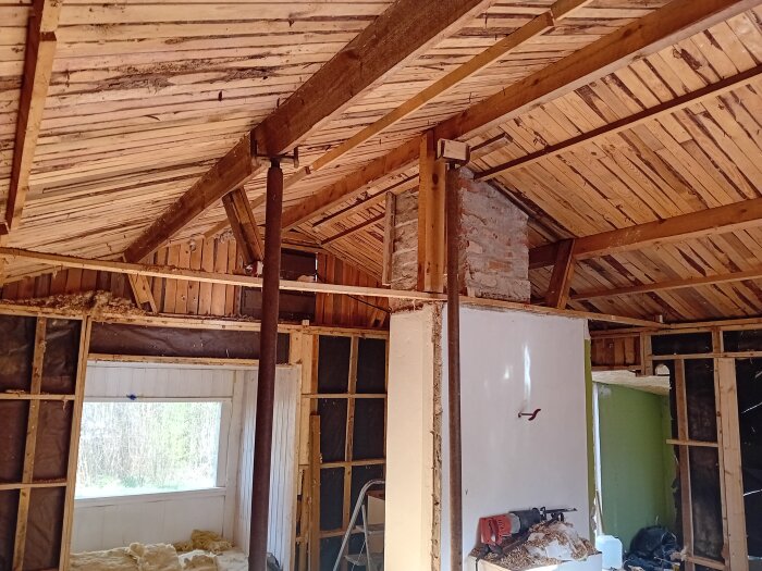 Inre delen av ett hus under renovering med synliga takbjälkar, murstock, och delar av väggar borttagna, samt byggmaterial runtom.