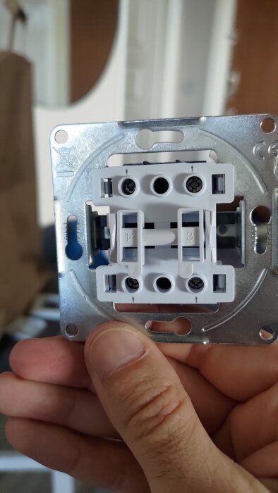 Framsidan av en strömbrytare, hålls i handen, visar skruvhål och anslutningspunkter.