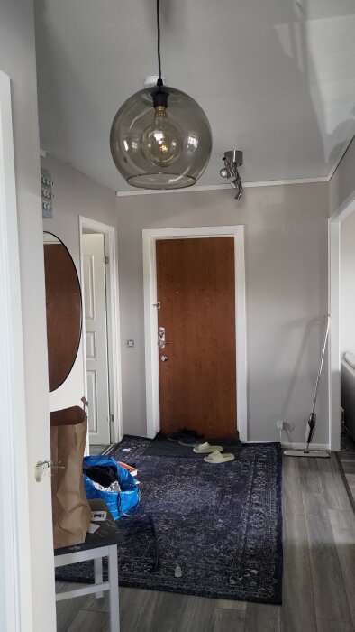 Entréhall med hängande glastaklampa och spotlights i taket, dörrmatta på golvet, en rund spegel och en öppnad dörr.