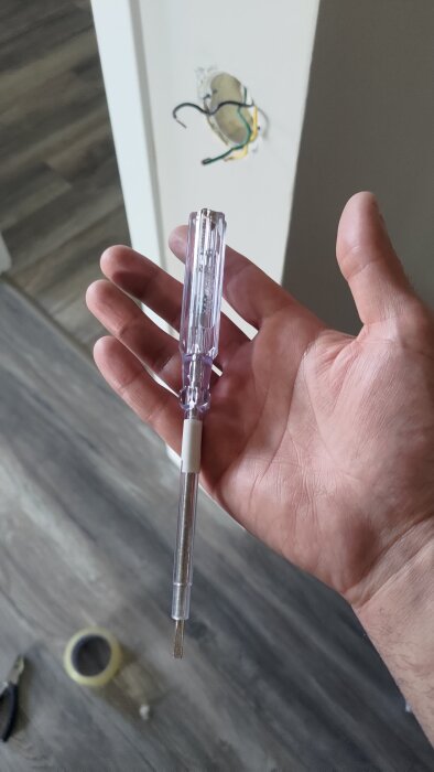 En hand håller i en spänningsprovare framför vägg med synliga svarta, gröna och gula elkablar.