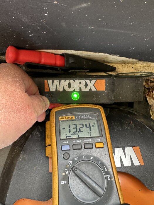 Multimeter visar 13,24V vid en Worx laddningsstation med grön lampa.