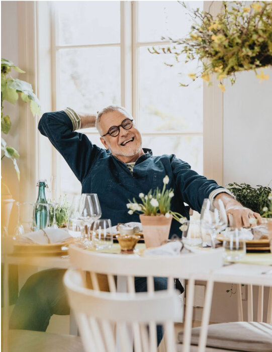 Man sitter avslappnat vid ett dukat bord med blommor och glas, ler och önskar glad påsk.