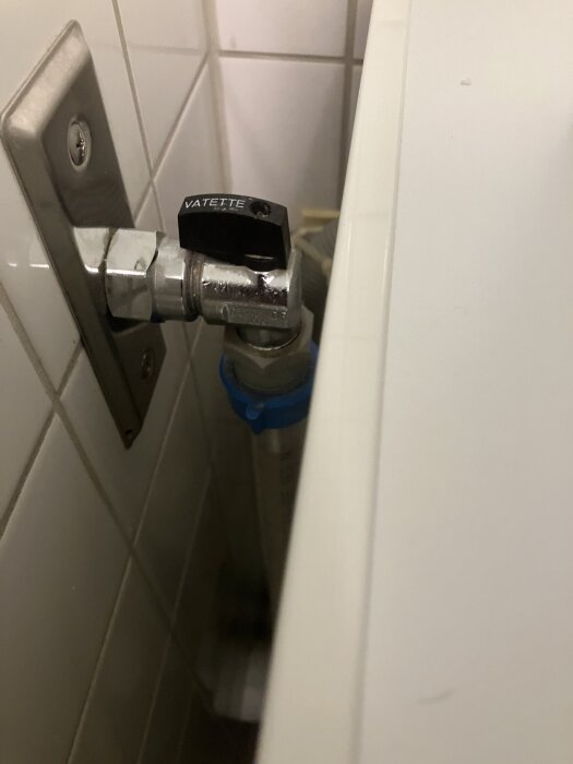 Kulventil med etiketten "VATETTE" installerad bakom tvättmaskin, fastsatt vid kakelfodrad vägg.