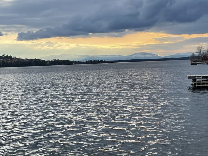 Vy över en sjö med högt vattenstånd och snöklädda fjäll i bakgrunden, sedd från en brygga i solnedgången.