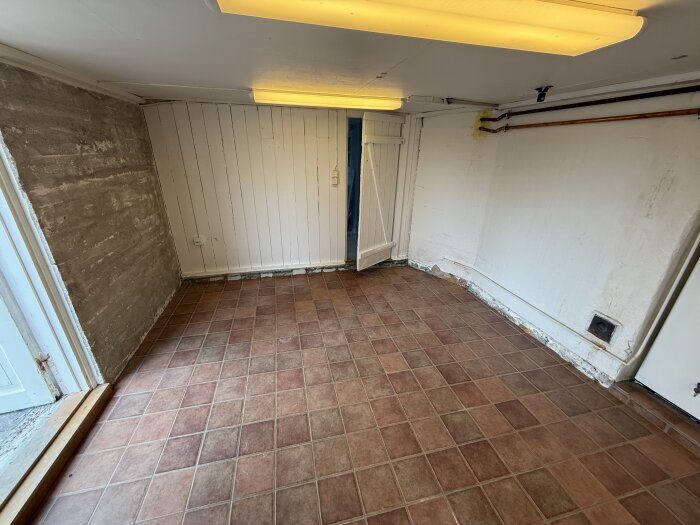 Rum med klinkergolv, vitmålad panel på vägg och synliga rörledningar.