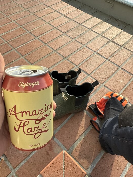 En hand håller en burk Stigbergets Amazing Haze IPA 6,5% på en terrass med golv av tegelstenar, bredvid stövlar och arbetshandskar.