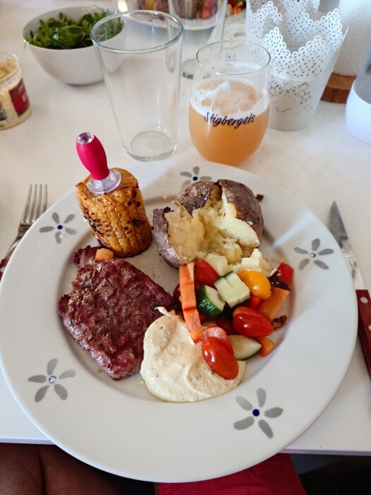 Grillad entrecote med bakad potatis, majs, grönsaker och sås på en tallrik.