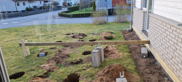 Konstruktion av altandäck vid en husvägg, med träbalkar och jordgropar för grundläggning, omgiven av en gräsmatta i ett bostadsområde.