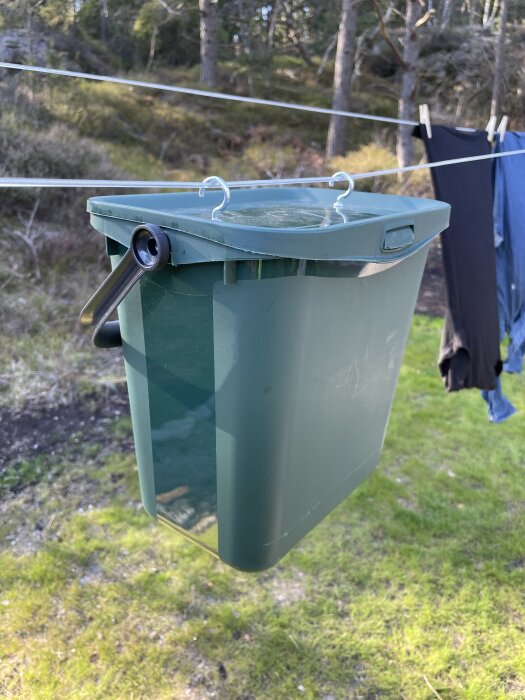 Grön återvinningshink med lock och krokar hänger på en tvättlina utomhus, med kläder i bakgrunden.