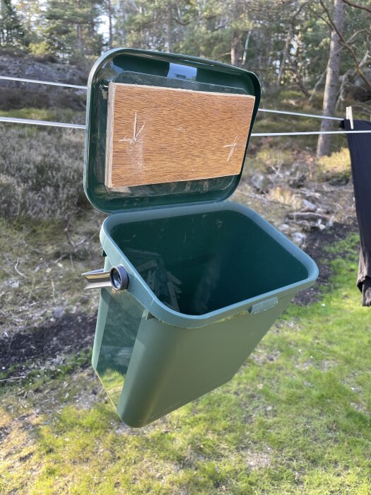 Grön återvinningshink från Biltema med lock och träbit inuti, hängandes på lina utomhus för förvaring av klädnypor.