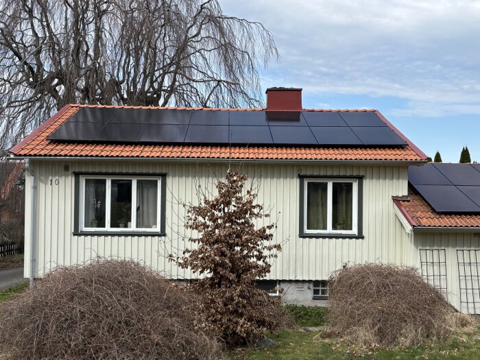 Småhus med vit träfasad och solpaneler på rött tak, omgiven av torra buskar och träd, under mulen himmel.