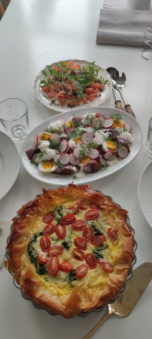Påsklunch med smördegsquiche med tomater, lax med kapris och rödbetssallad med ägg och rädisor på ett vitt bord.