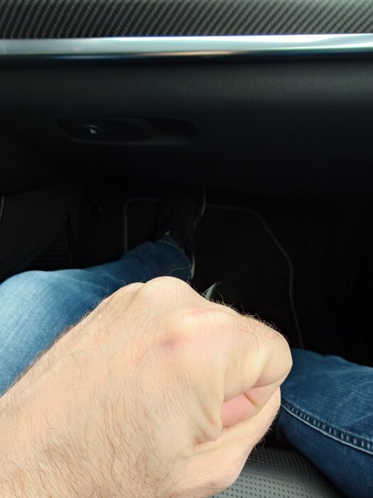 En knuten näve syns framför en ben iklädd jeans i en bilinteriör.