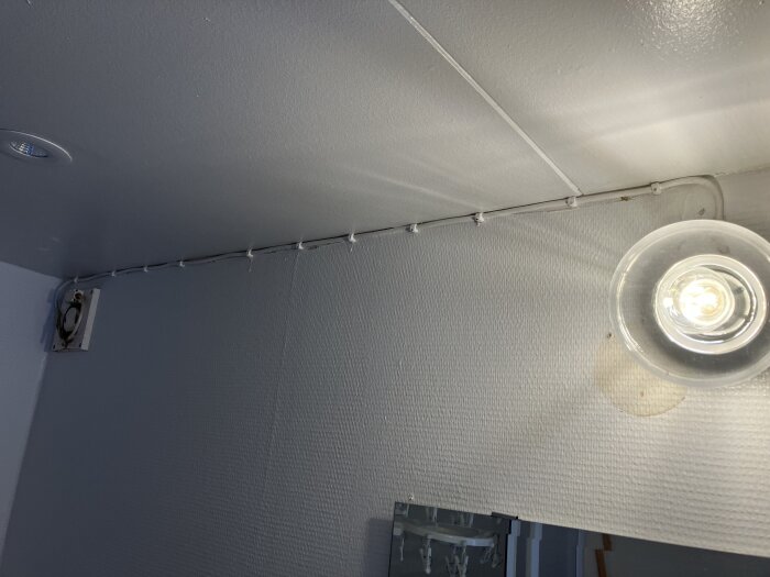 En fläkt monterad på väggen i ett rum, kopplad till armaturen, med sladd fäst längs taket.