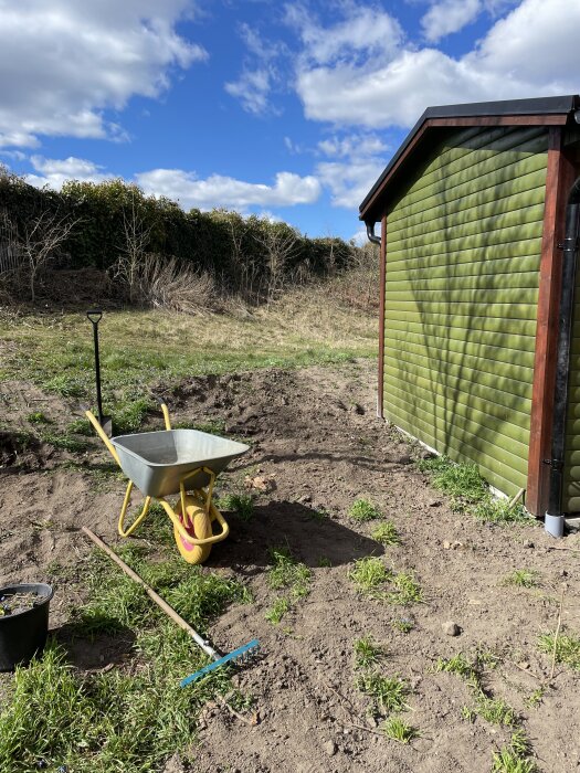 Skottkärra och redskap vid en grön byggnad på en jordig tomt med blå himmel i bakgrunden.