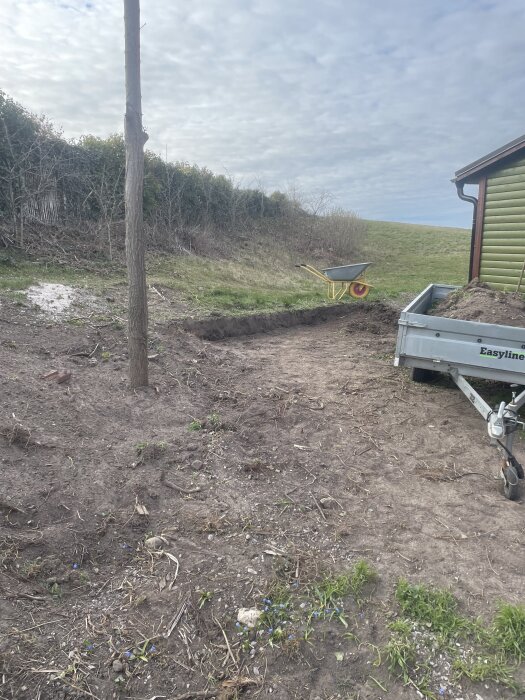 Grävning för nivåmur med skottkärra och trailer, bakomliggande gräsmatta och häck.