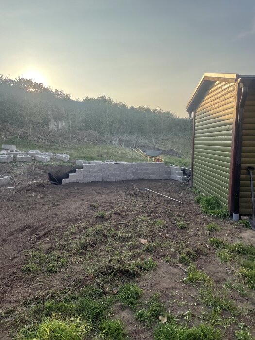 Mur av grå betongblock bredvid ett grönt hus, omgiven av jord och gräs, med en skottkärra och kvällssol i bakgrunden.