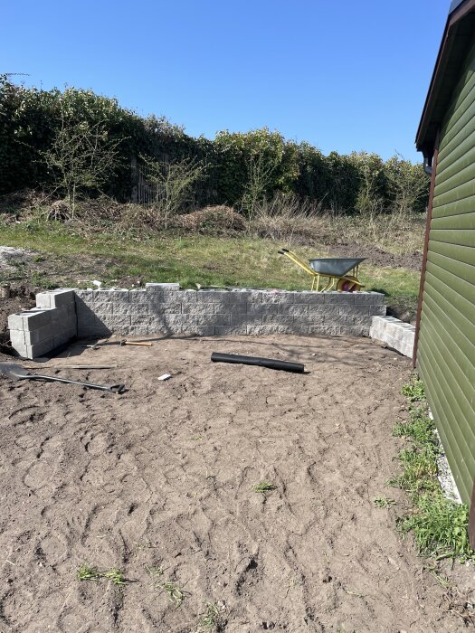 Nivåmur under konstruktion i trädgård med verktyg och skottkärra; jordområde runtom.