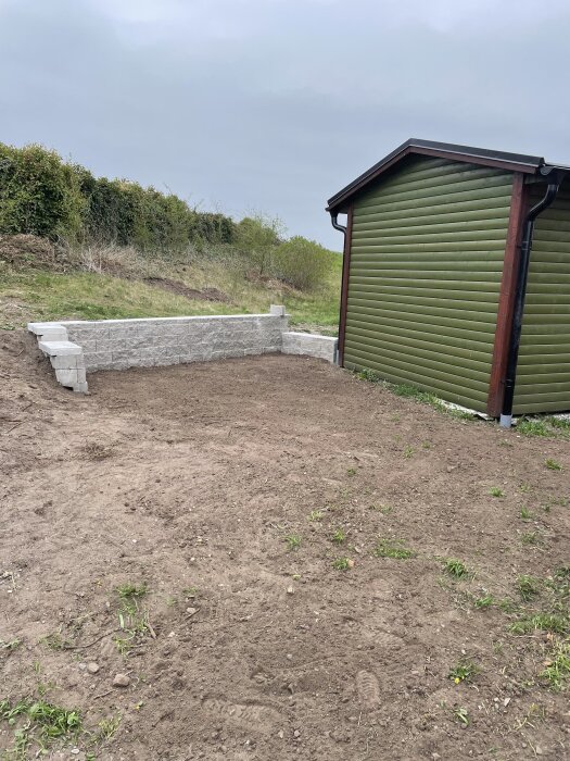 Nivå mur av vit sten framför en grön byggnad vid kanten av en tomt, med nyligen omfördelad jord runt omkring.