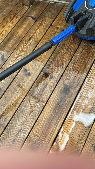 Trädäck med en högtryckstvätt i färd med att rengöra ytan. Det finns rester av skum och våta partier på träet.