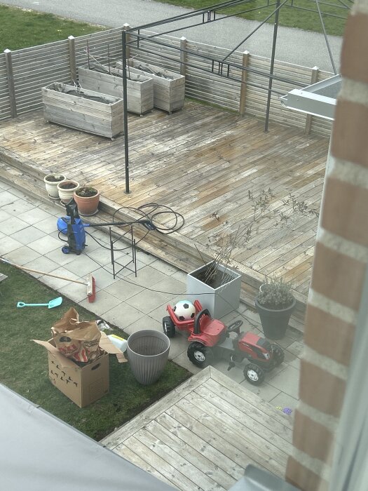 Vy över trädäck med planteringslådor, leksakstraktor och fotboll, sett uppifrån. Trädäcket längst ned är behandlat med såpa och tralltvätt.