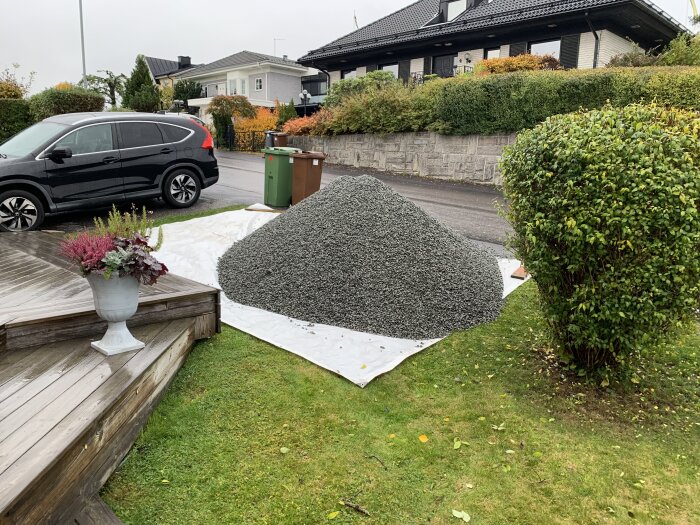Hög av makadam på gräsmatta bredvid svart bil och trädgård, täckt av vit presenning.
