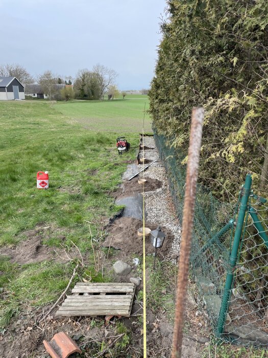 Borrade gjutrör och ett gammalt staket syns vid en buske längs med ett grönt fält, med verktyg och material för renovering liggande på marken.