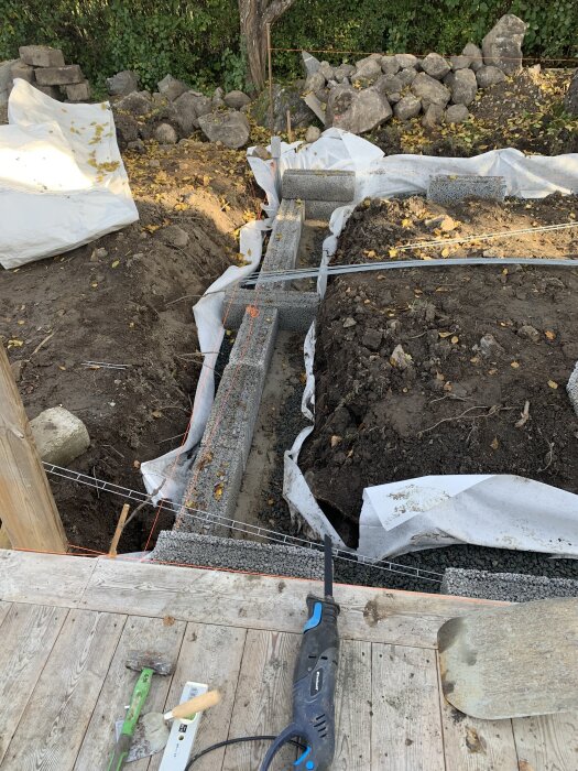 En byggarbetsplats med murstenar som formar en stödmur på lutande mark, med betongsula, verktyg och jordlager.