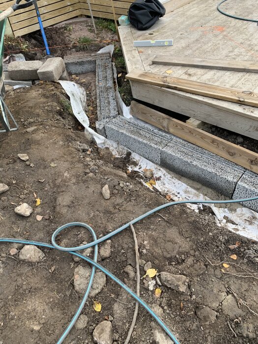 Murstenar och träplankor vid en utgrävd plats med betongsula. Verktyg ligger på träplattformen i bakgrunden.